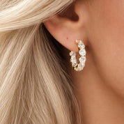 Close-up of a gold hoop earring with clear stones worn by a person with blonde hair.