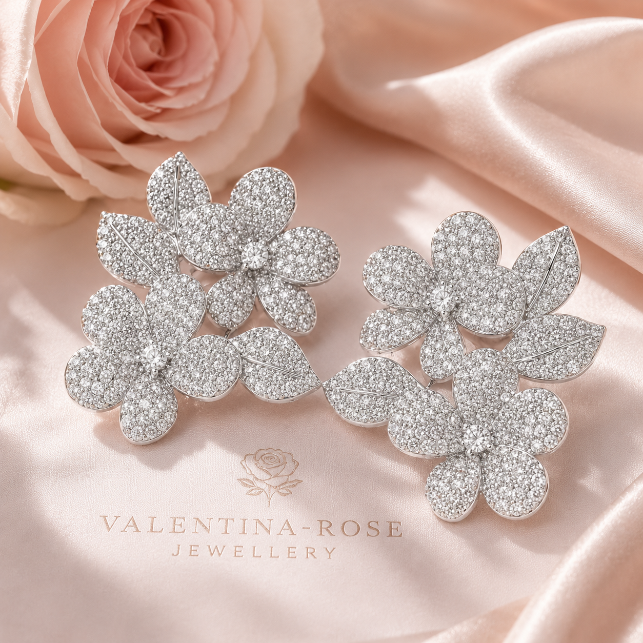 A pair of silver-toned flower-shaped stud earrings covered in clear pavé stones, displayed on soft pink satin fabric with a pink rose in the background. The brand name 'VALENTINA-ROSE JEWELLERY' is printed on the fabric below.