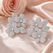 A pair of silver-toned flower-shaped stud earrings covered in clear pavé stones, displayed on soft pink satin fabric with a pink rose in the background. The brand name 'VALENTINA-ROSE JEWELLERY' is printed on the fabric below.