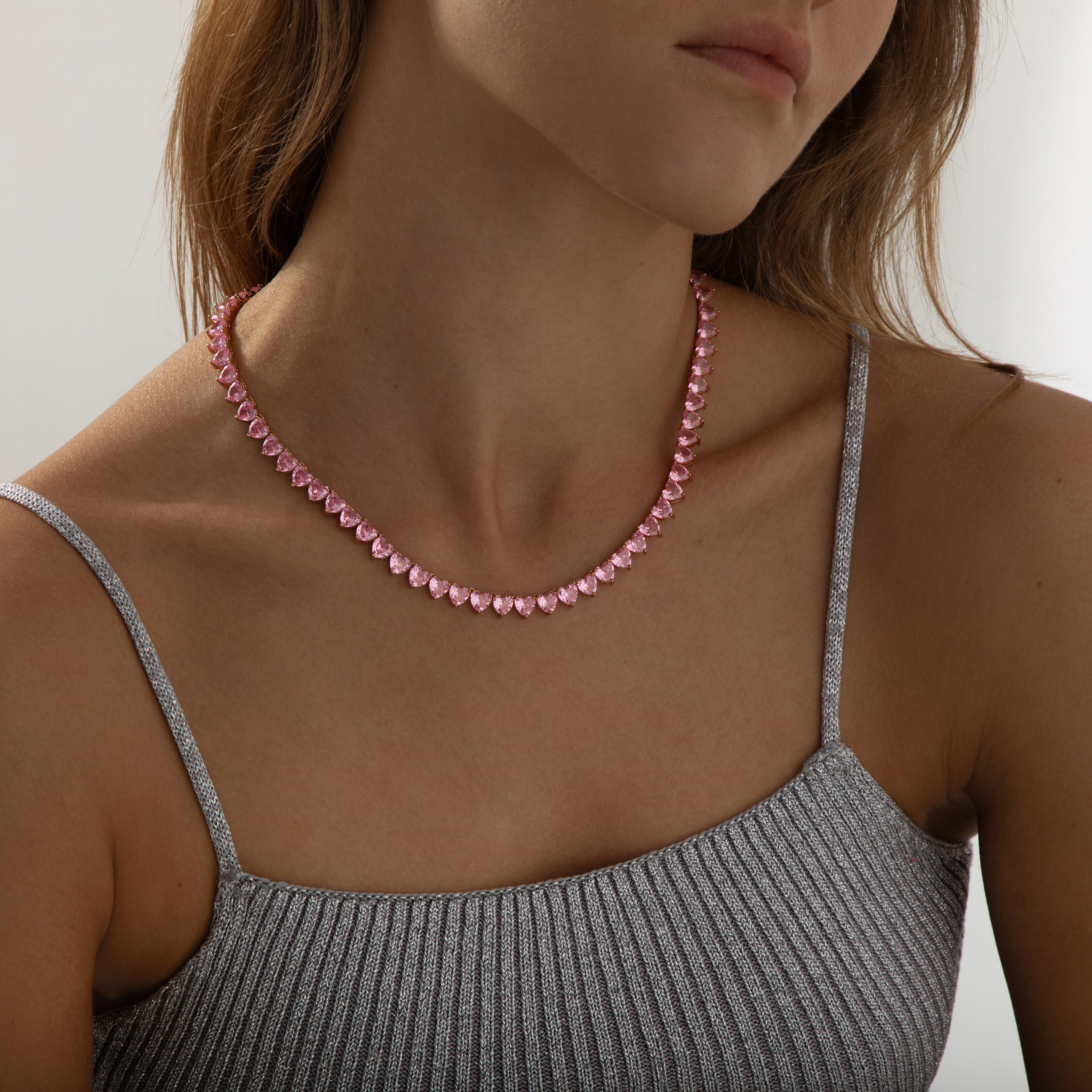 A rose‑gold plated tennis‑style necklace drapes elegantly around a model’s neck. The piece features a continuous row of blush‑pink, heart‑shaped cubic zirconia stones, each approximately 5 mm wide, linked closely together. The necklace measures about 17 in (43 cm) with a 0.5 in extender and fastens with a secure clasp. The polished rose‑gold setting enhances the sparkle of the pink hearts, creating a romantic, feminine shimmer perfect for both everyday wear and special occasions. 