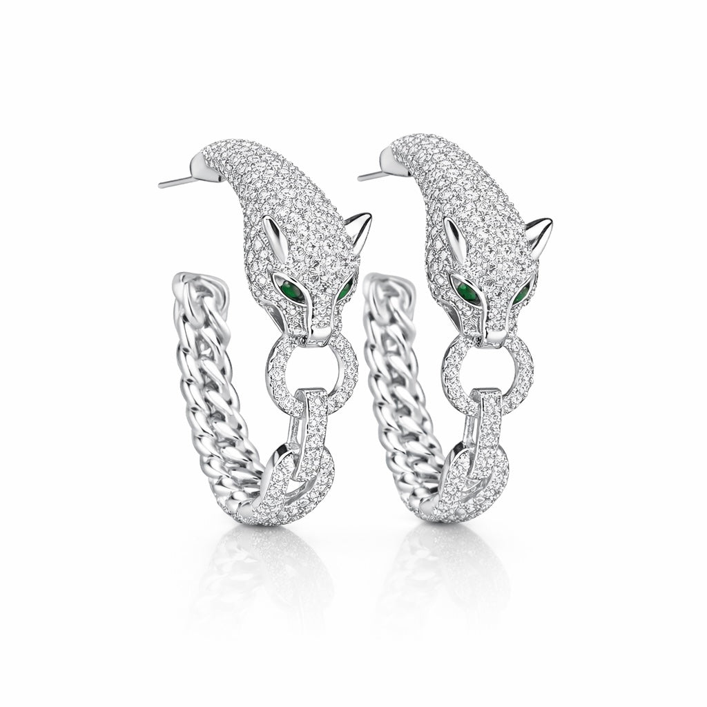 A pair of silver hoop earrings featuring a detailed panther head design paved with clear stones, with small green emerald accents in the eyes, shown on a white background.