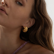 A pair of chunky, semi-circular hoop earrings photographed on a model. Each hoop is crafted from stainless steel plated with glossy 18K gold, featuring an embossed design of small buttercup flowers densely covering the outer surface. The hoops are approximately 2.4 cm tall and 1.8 cm wide, with a smooth, reflective finish. They weigh around 15 g and appear waterproof and tarnish-resistant, making them an everyday-ready, statement floral accessory.
