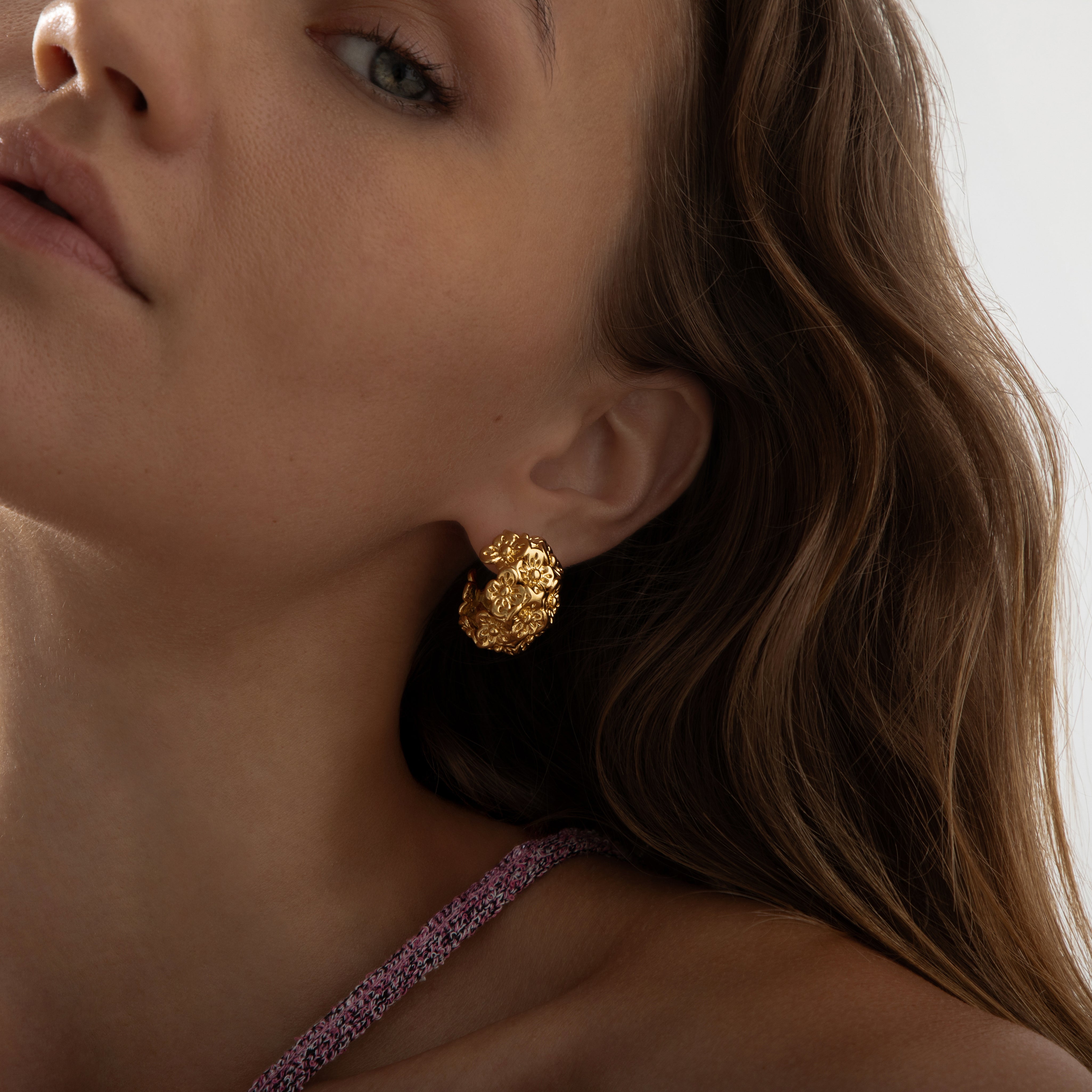 A pair of chunky, semi-circular hoop earrings photographed on a model. Each hoop is crafted from stainless steel plated with glossy 18K gold, featuring an embossed design of small buttercup flowers densely covering the outer surface. The hoops are approximately 2.4 cm tall and 1.8 cm wide, with a smooth, reflective finish. They weigh around 15 g and appear waterproof and tarnish-resistant, making them an everyday-ready, statement floral accessory.
