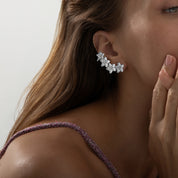 
A pair of rhodium-plated ear climbers arranged symmetrically on a white background. Each earring features a graceful, curved line of four stylised flower motifs that follow the ear’s contour. The flowers are fully pavé-set with round cubic zirconia crystals, with one slightly larger bloom at the bottom transitioning to smaller blossoms toward the top. The climbers measure approximately 4.1 cm in height and 2.3 cm in width, and are secured via a long post at the base and a secondary backing near the top to 