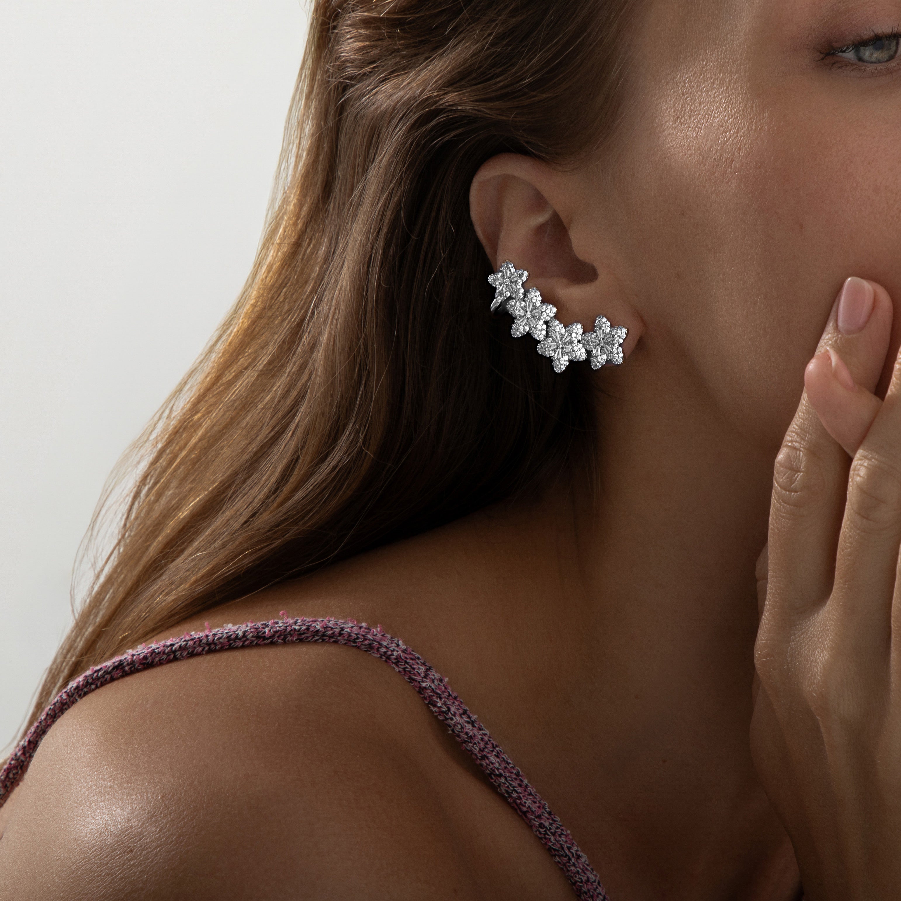 
A pair of rhodium-plated ear climbers arranged symmetrically on a white background. Each earring features a graceful, curved line of four stylised flower motifs that follow the ear’s contour. The flowers are fully pavé-set with round cubic zirconia crystals, with one slightly larger bloom at the bottom transitioning to smaller blossoms toward the top. The climbers measure approximately 4.1 cm in height and 2.3 cm in width, and are secured via a long post at the base and a secondary backing near the top to 