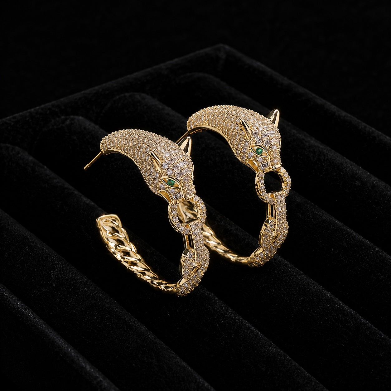 A pair of gold hoop earrings designed as stylized panther heads, heavily embellished with small clear stones and featuring tiny green gemstone eyes, displayed on a black velvet jewelry box.