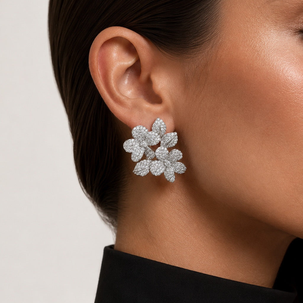 A close-up of a model's ear wearing a silver-toned flower-shaped stud earring paved with small clear stones, against a light neutral background.