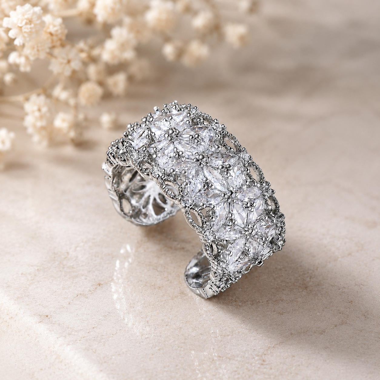 A wide silver cuff ring embellished with clear pavé-style stones, photographed on a light beige stone surface with soft cream flowers in the background.