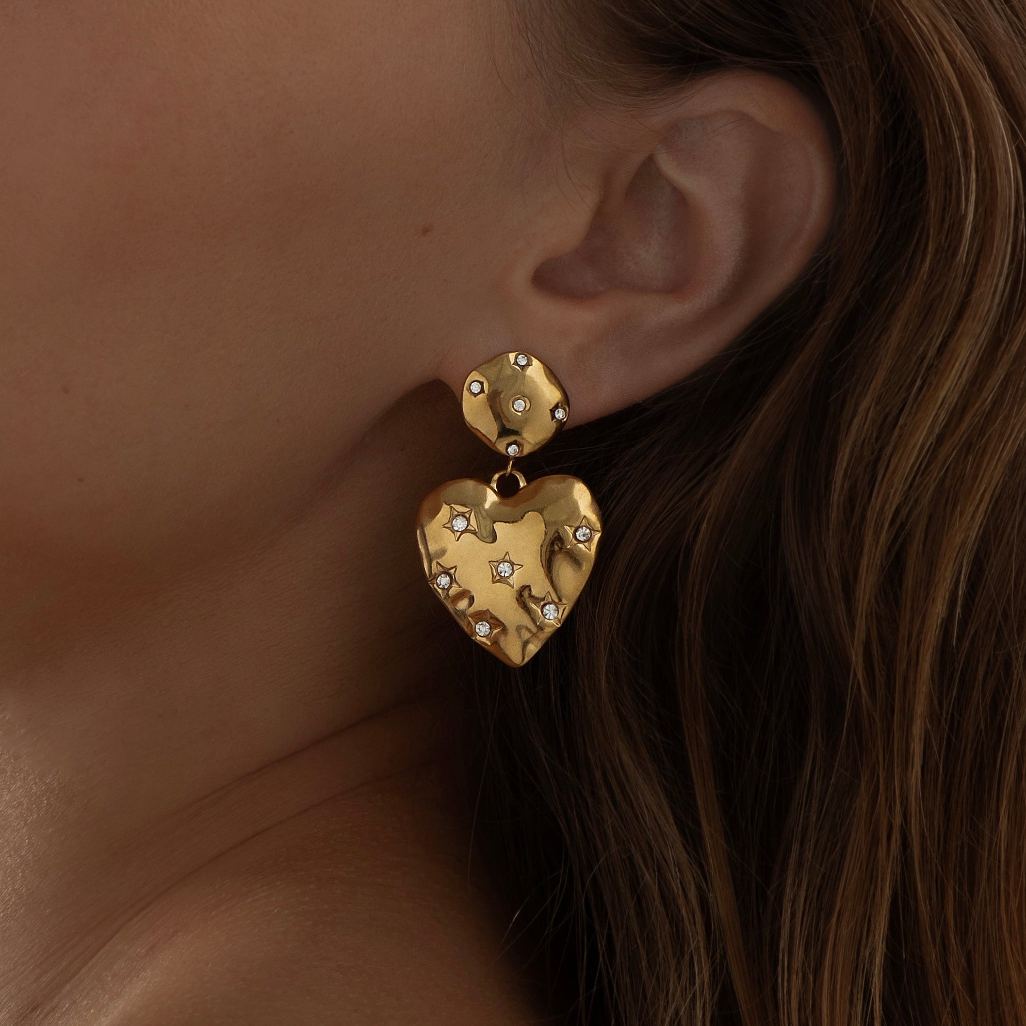 Gold heart-shaped earring with embedded stones worn by a person.
